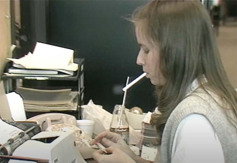 A screenshot from a 1985 CBC news segment on cigarette smoking in the workplace, showing a young woman smoking in an office at a typewriter.