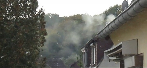 The side of a house. Across the way are trees, and trees can be seen in the background. A thick opaque cloud of smoke billows from a chimney on the roof of the house.