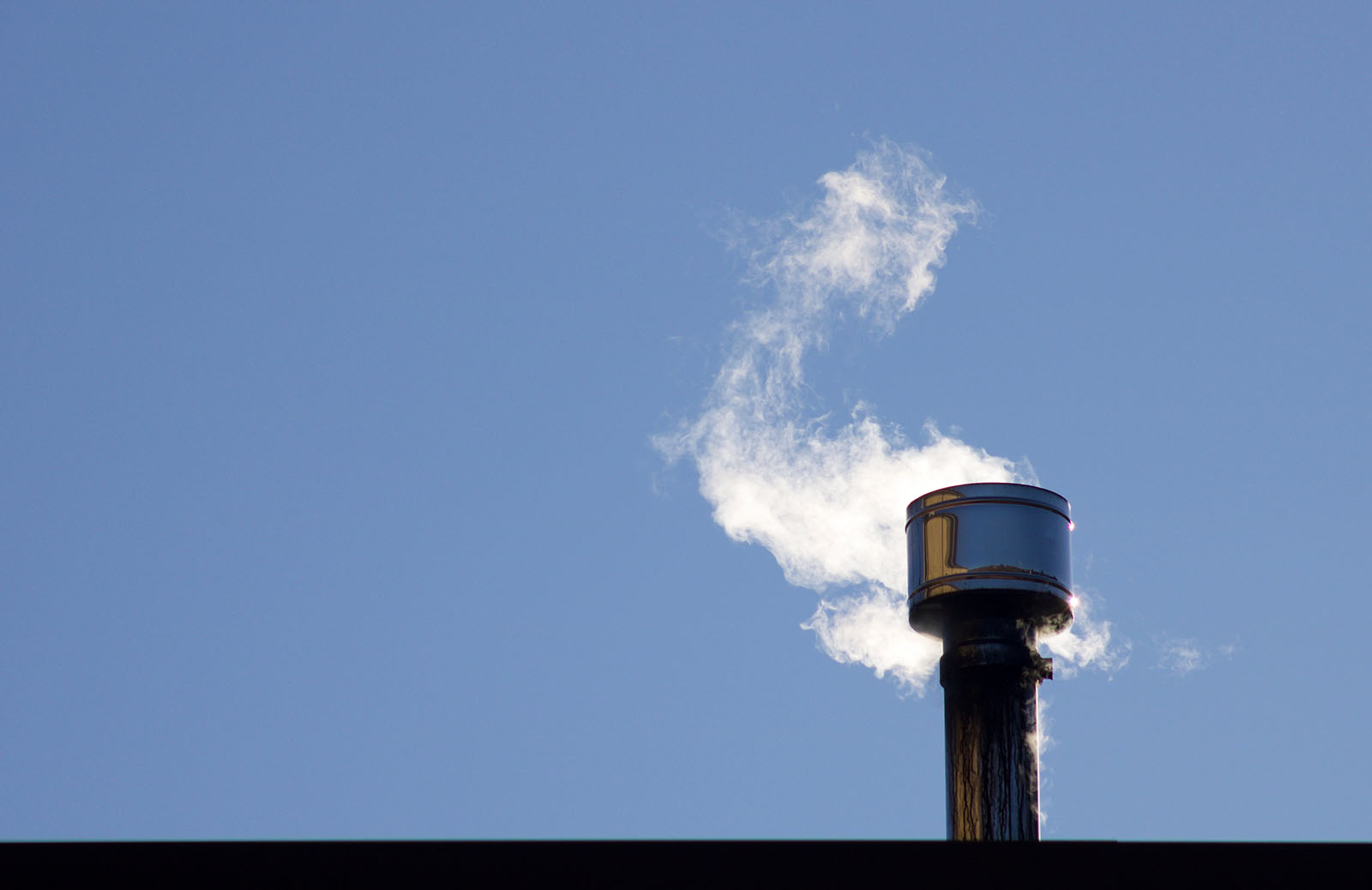 A wood stove chimney emits smoke against a clear blue sky.
