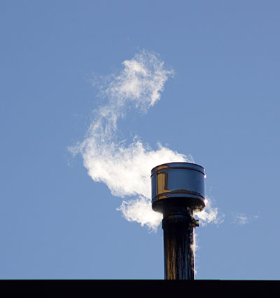 A wood stove chimney emits smoke against a clear blue sky.
