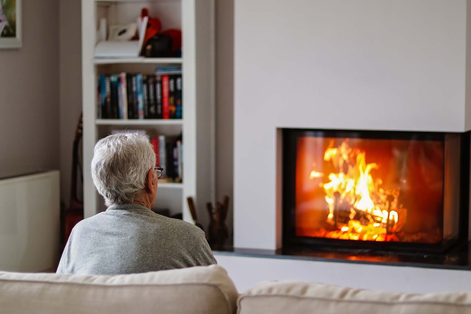An older man sits in his living room in front of a lit fire.