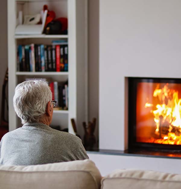 An older man sits in his living room in front of a lit fire.