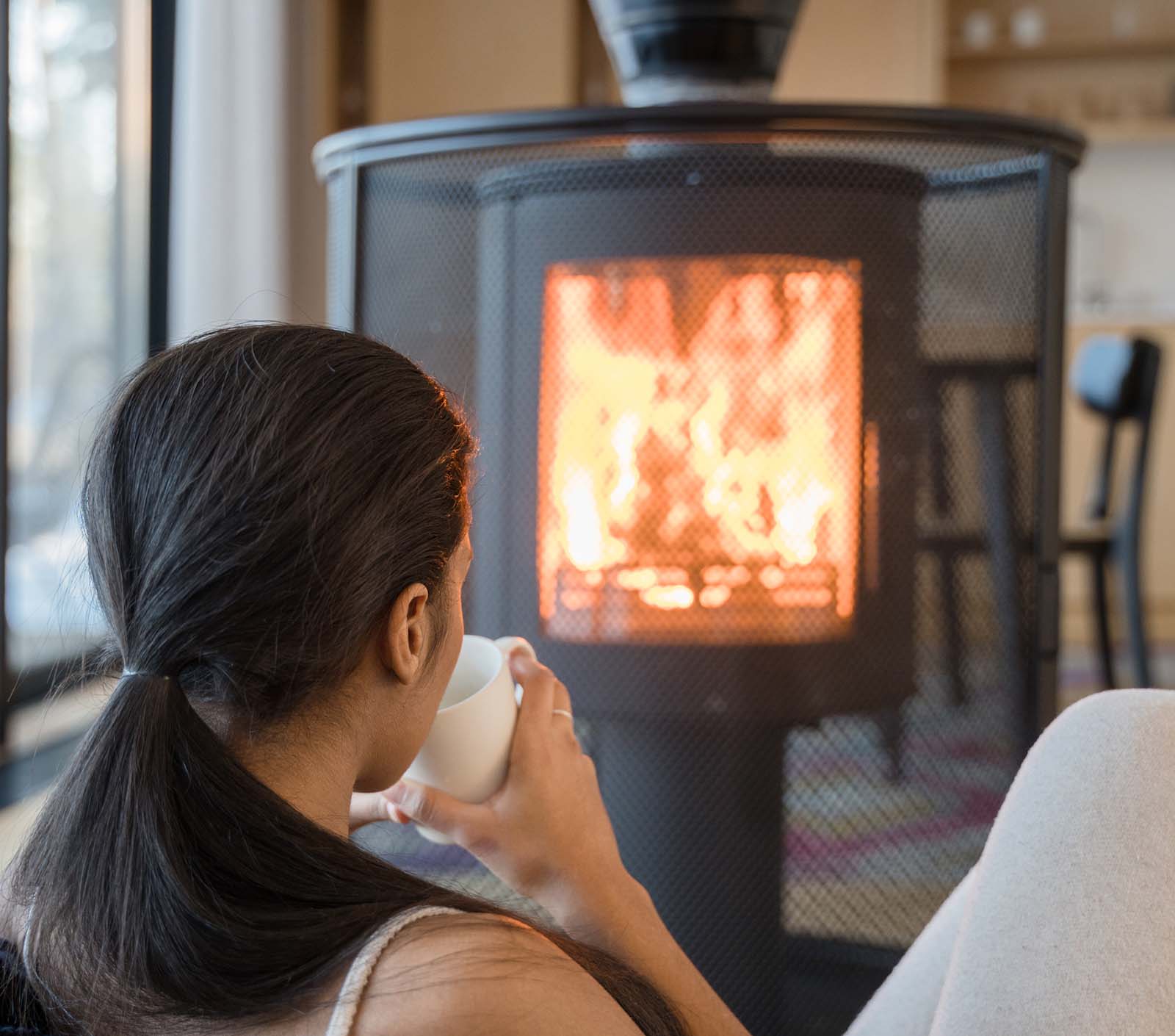 A woman sips from a mug in front of a lit wood stove.