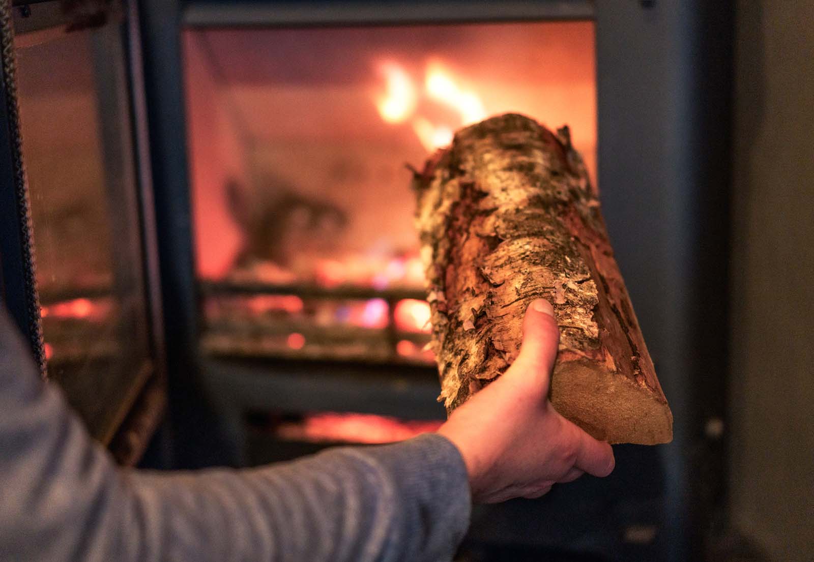 A closeup of someone putting a piece of wood into a lit wood stove.