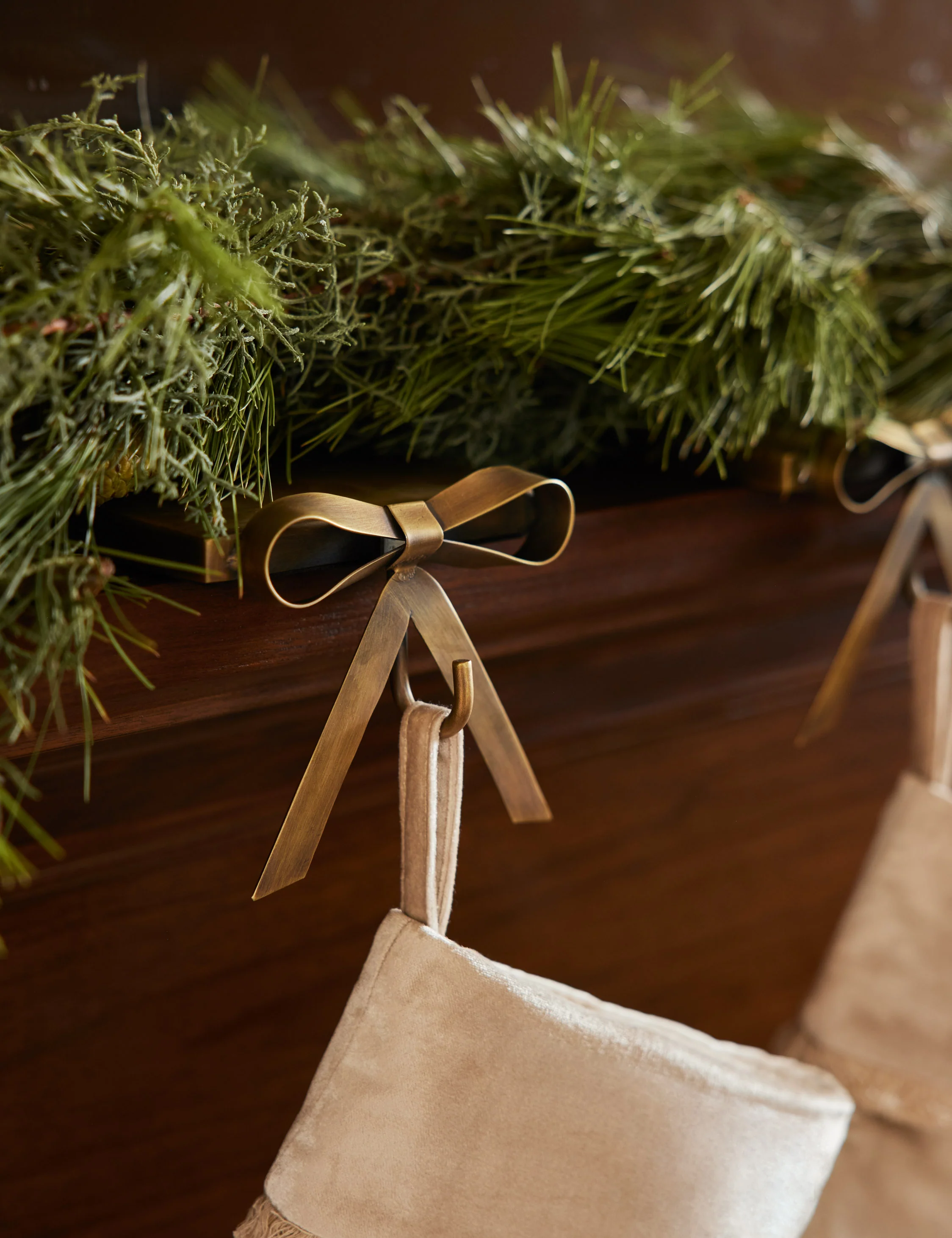Close-up of a beige Christmas stocking hanging from a brass bow-shaped hook on dark wood, with green garland on top.