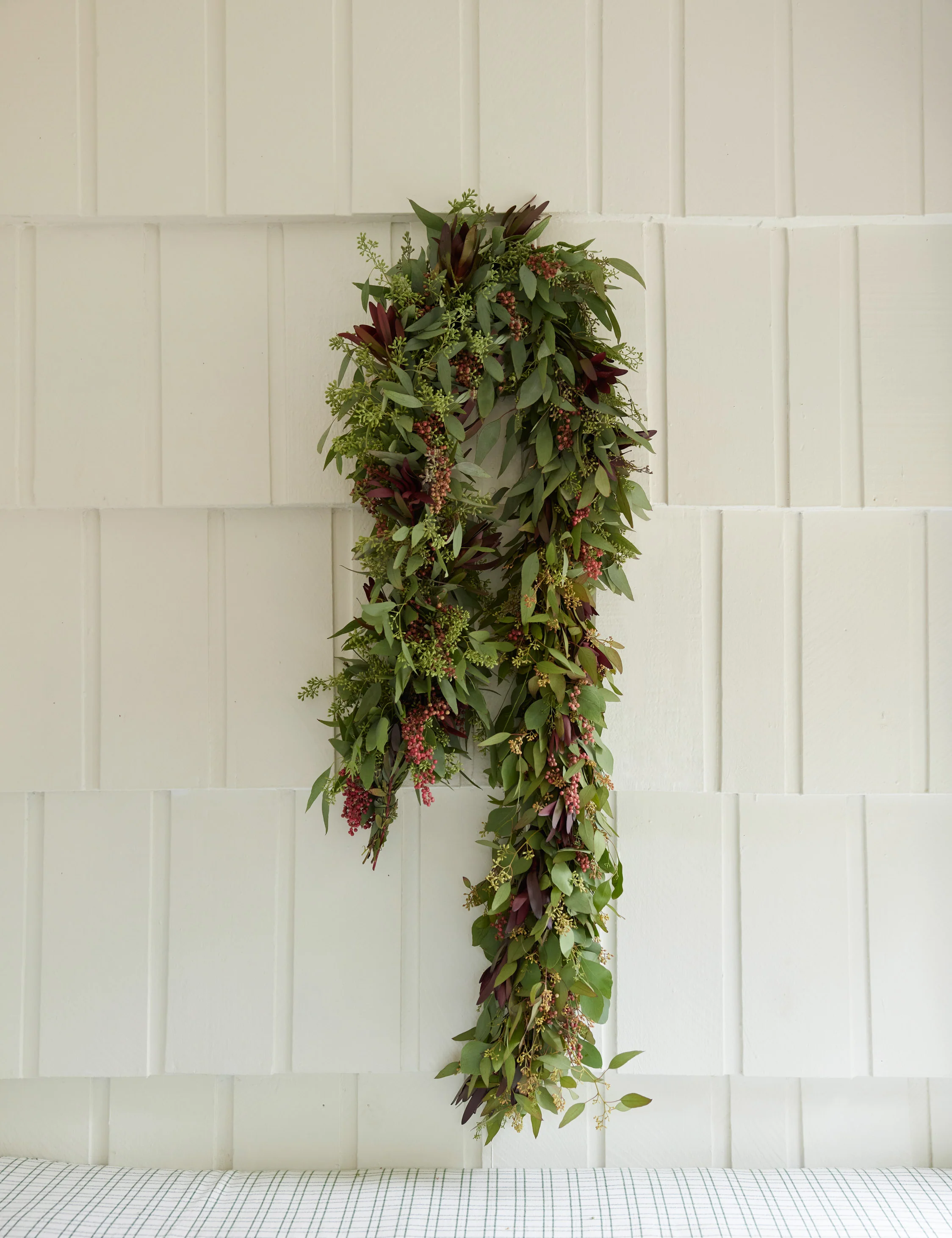 Green and burgundy leafy garland with small berries hanging on a white tiled wall above a checked cloth surface.