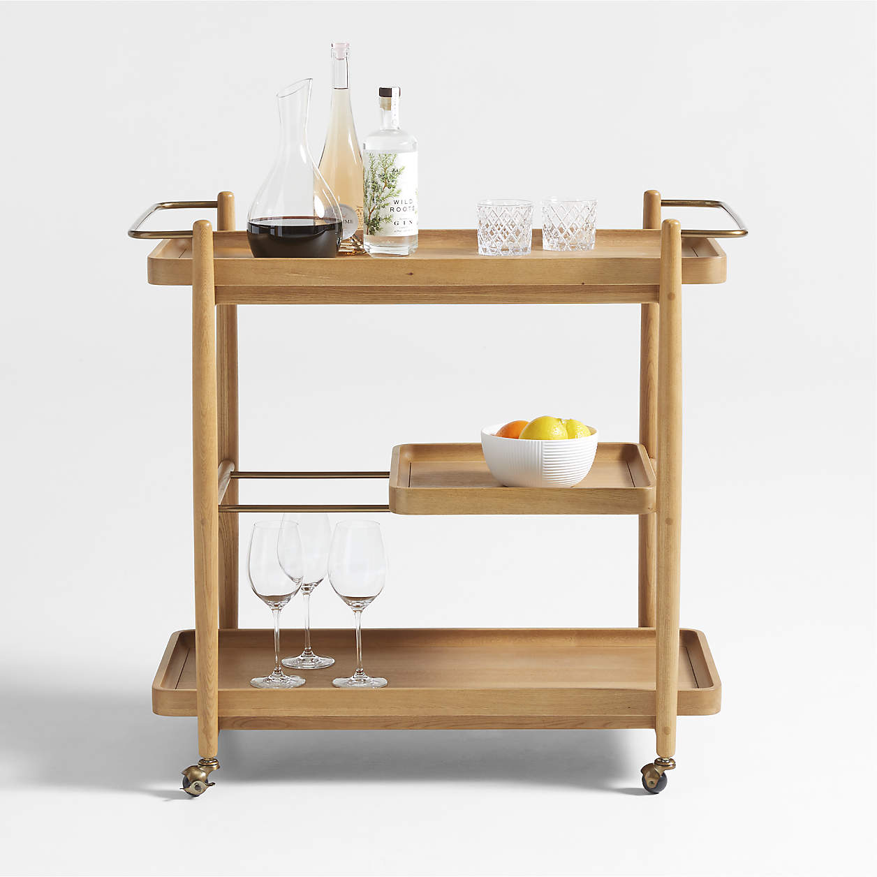 Wooden bar cart with wheels holding a decanter with red wine, two bottles of alcohol, two glass tumblers, a bowl of fruit, and three wine glasses.