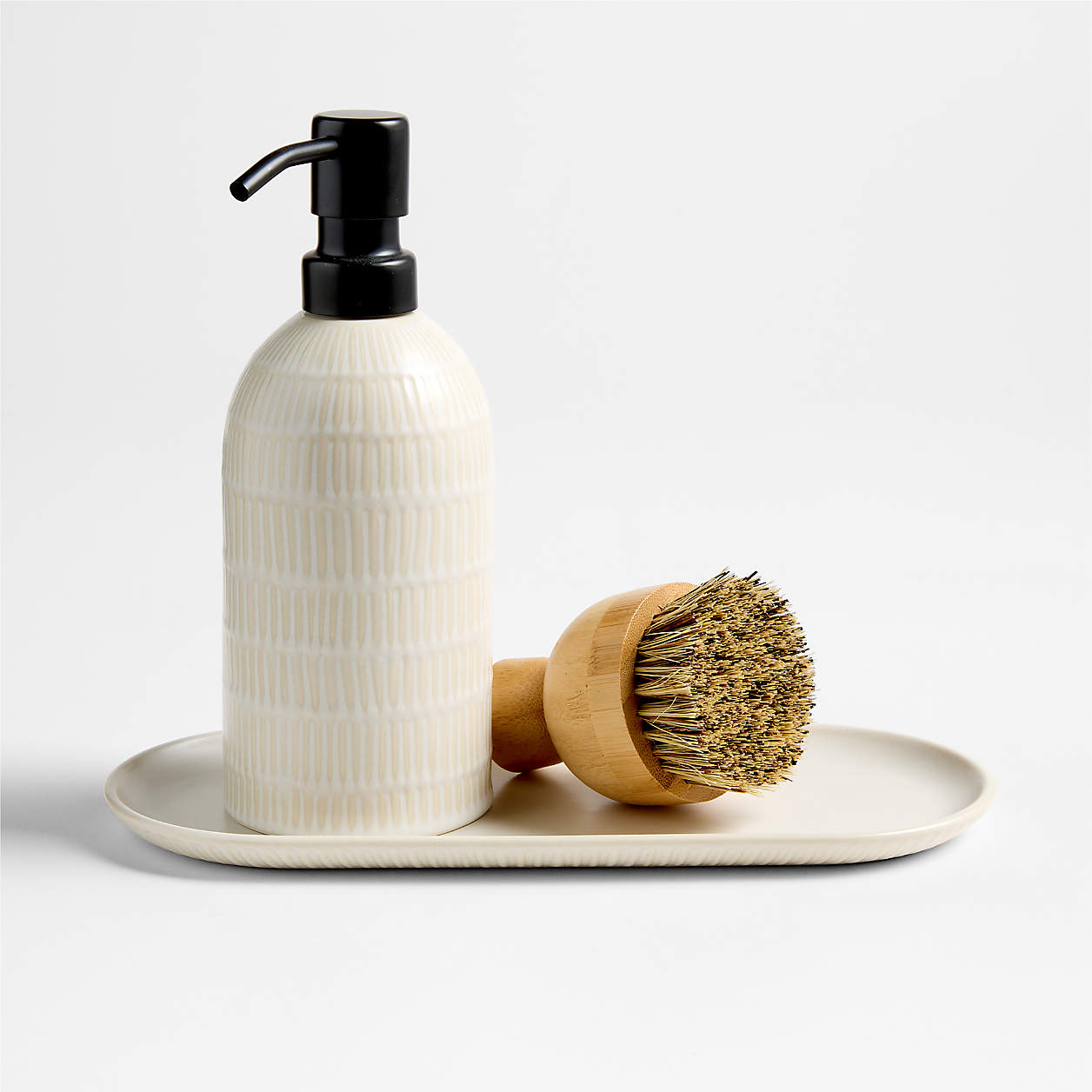 Ceramic soap dispenser with black pump and round wooden brush with stiff bristles on an oval beige tray.