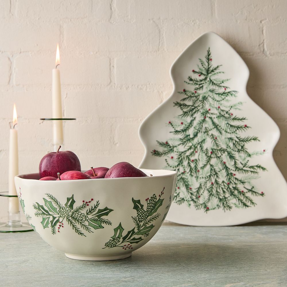 White ceramic bowl with green holly and pinecone design filled with red apples, in front of a white tree-shaped plate with a green pine branch pattern and two lit white candles in glass holders.