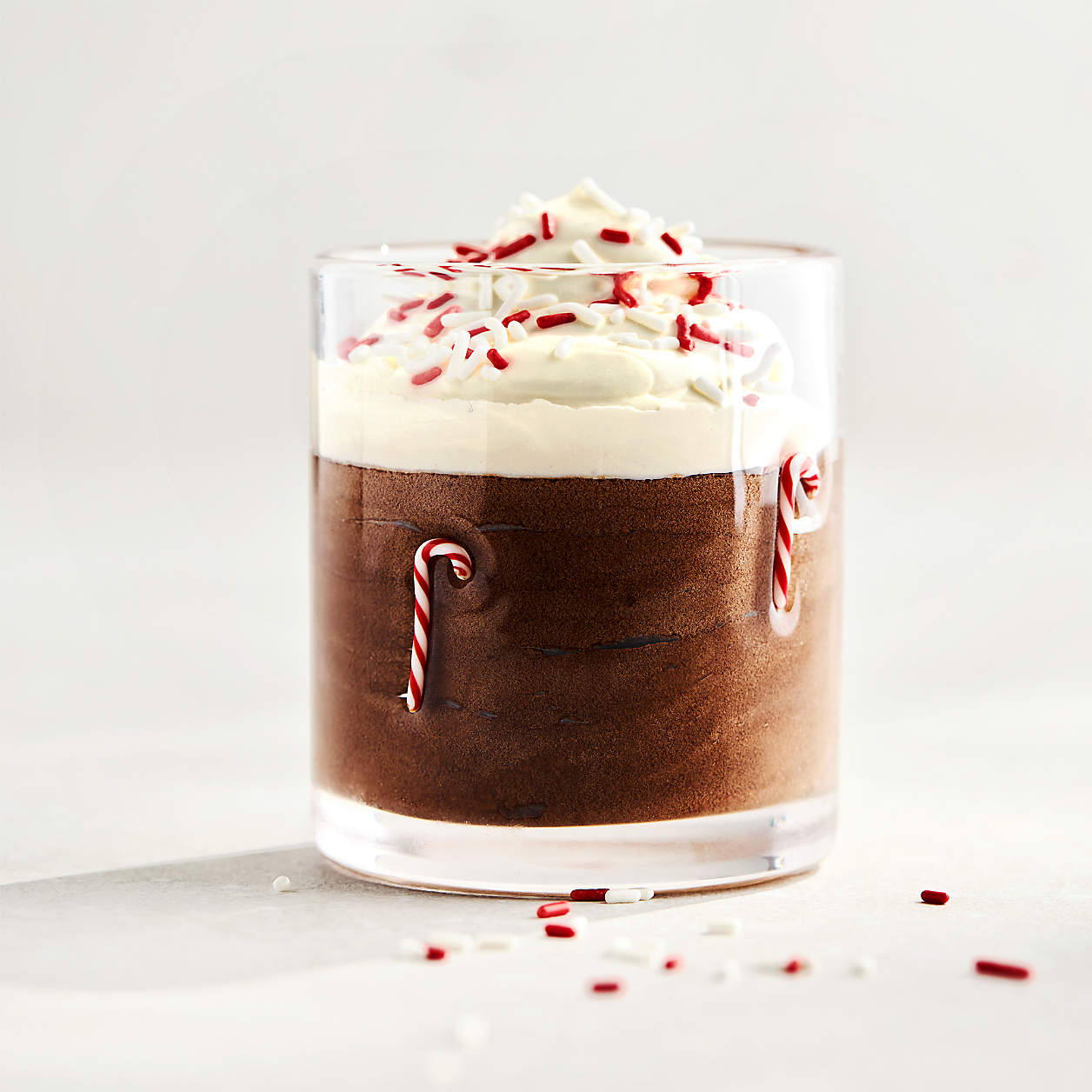 Clear glass filled with chocolate mousse topped with whipped cream and red and white sprinkles, decorated with small candy canes.