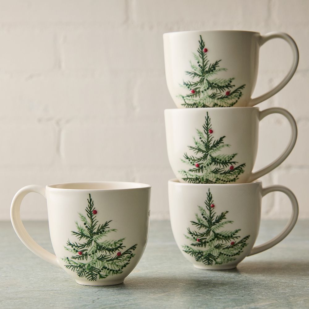Set of four white ceramic mugs decorated with green pine tree branches and red berries, three stacked and one beside them on a blue surface.