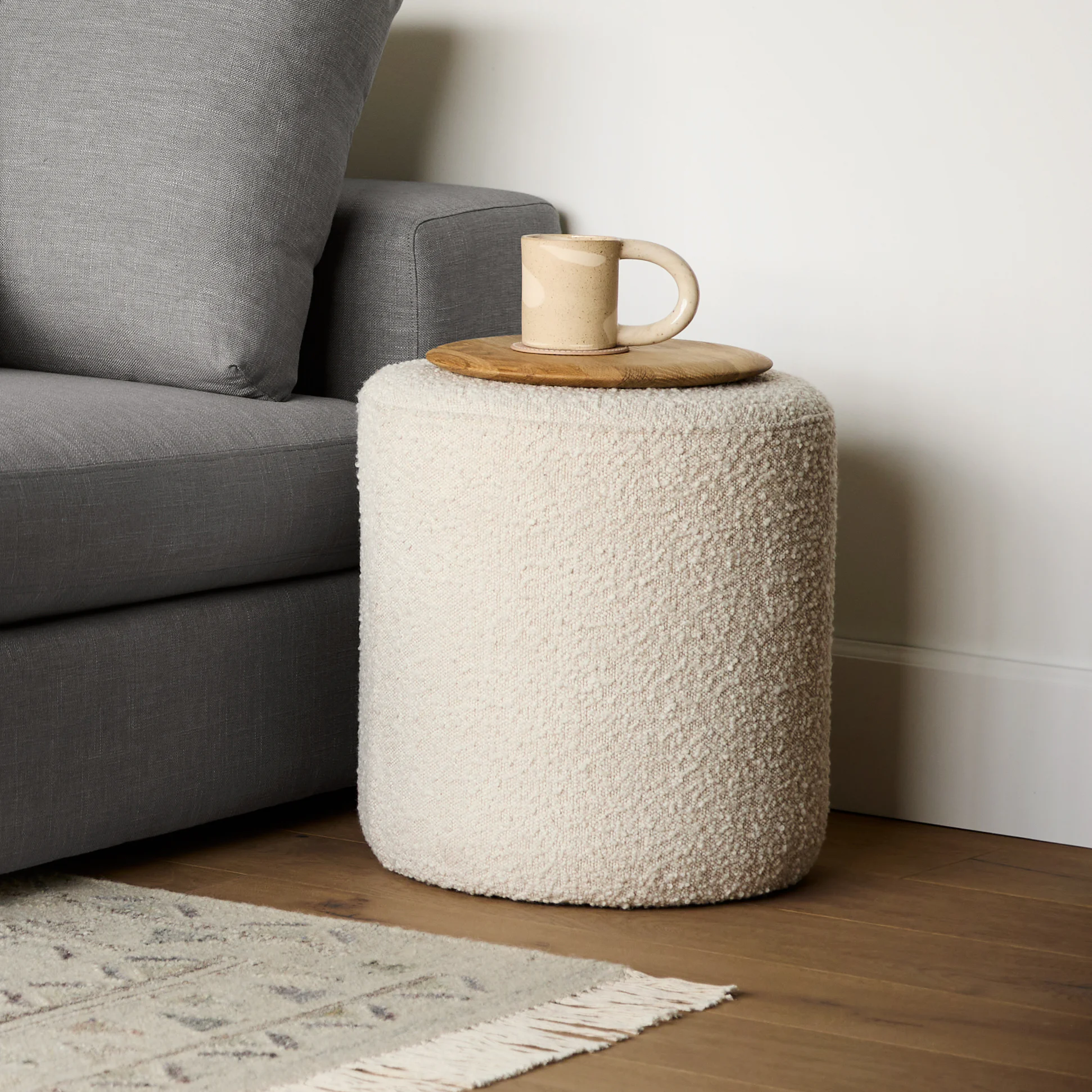 Beige textured cylindrical ottoman next to a gray sofa with a ceramic mug on a wooden tray on top.