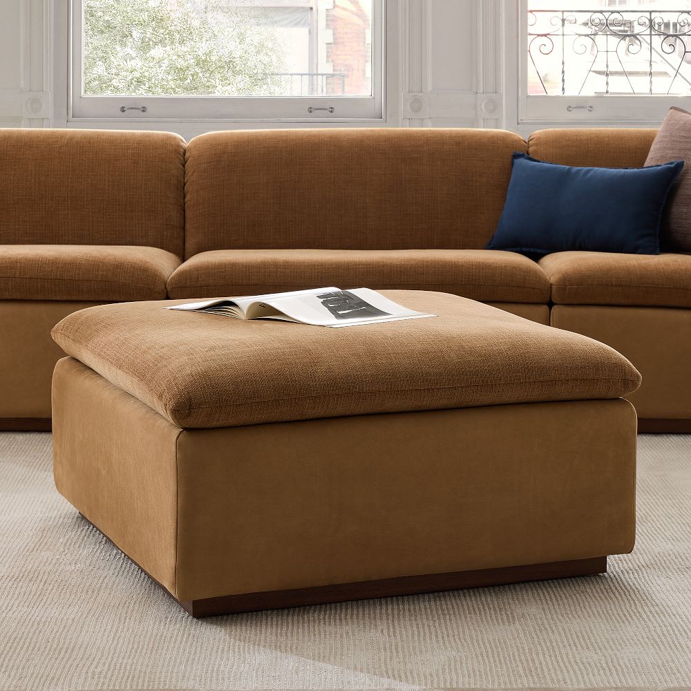 Brown fabric and leather ottoman with an open magazine on top, placed in front of a matching brown sectional sofa with a blue pillow.