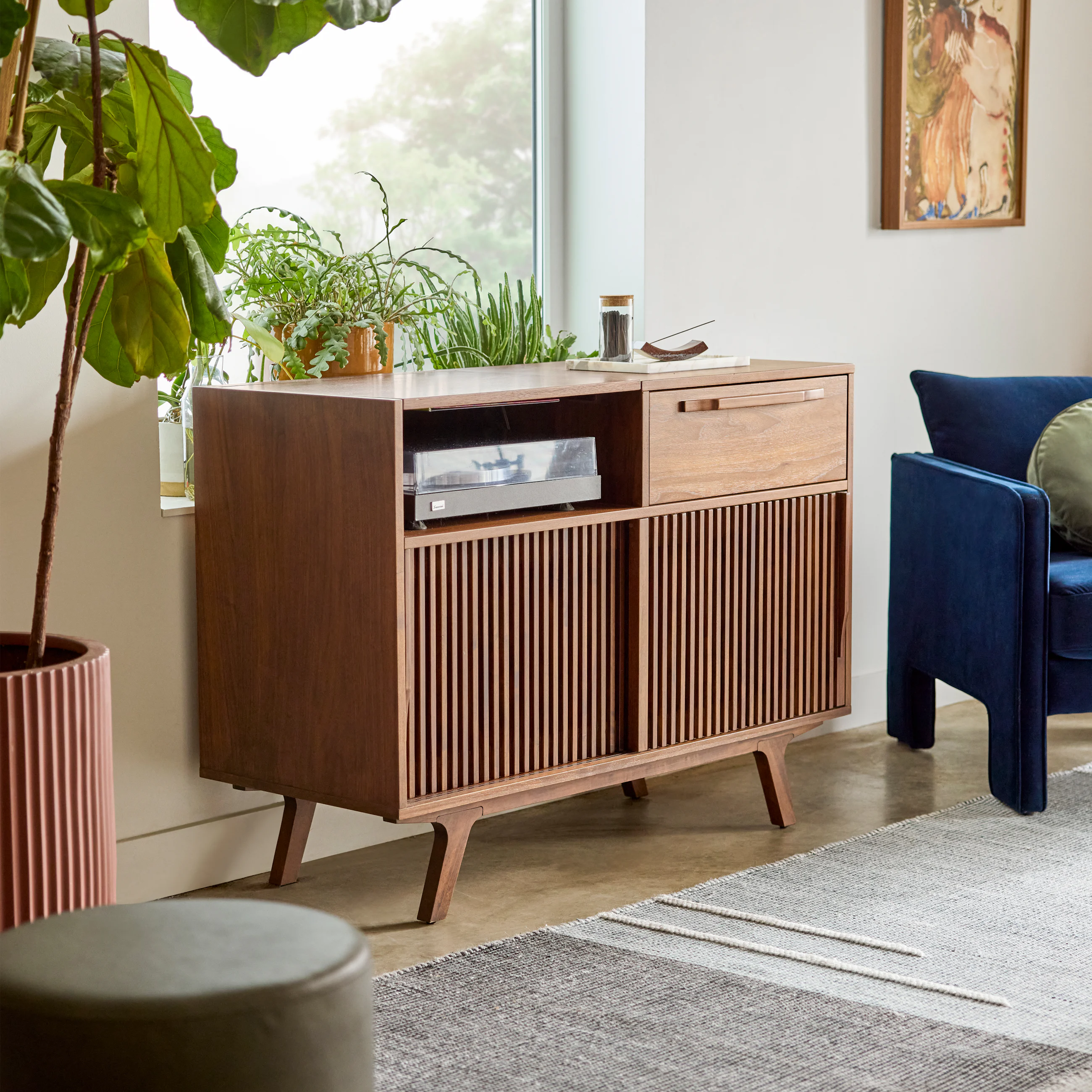 Mid-century modern wooden cabinet with a turntable, plants on top, next to a blue armchair in a bright living room.