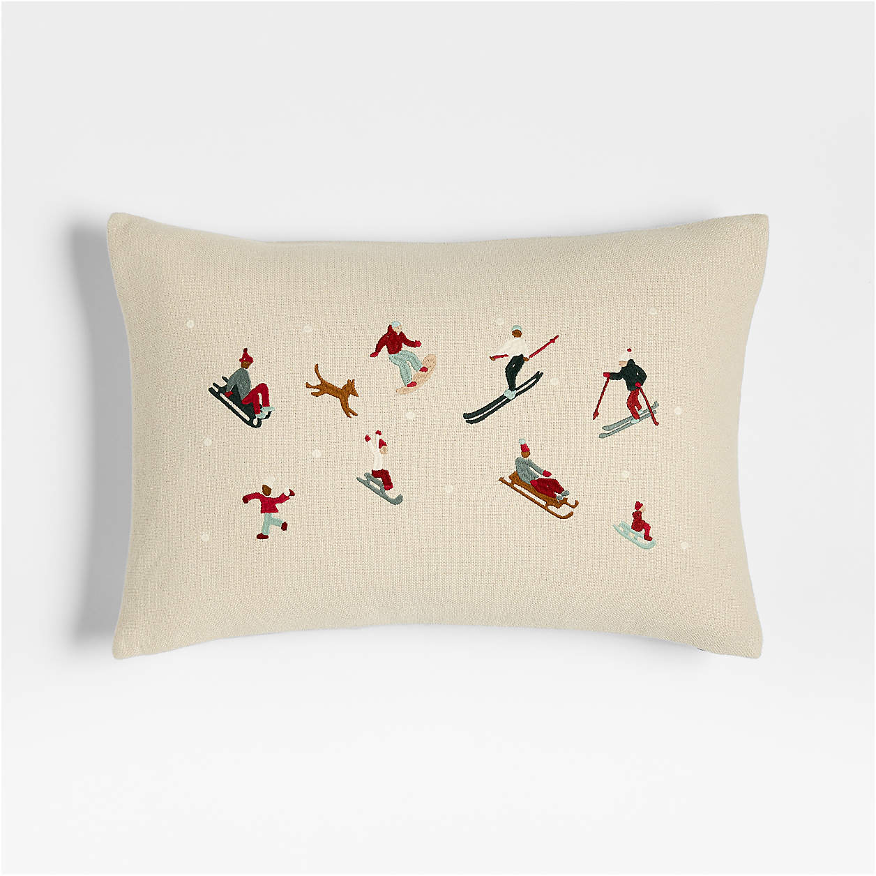 Beige rectangular pillow with embroidered winter scene of people skiing, sledding, snowboarding, and a dog playing in the snow.