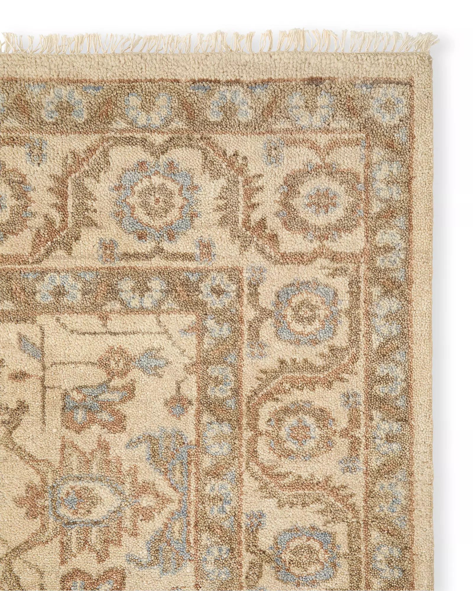 Corner of a beige wool rug with intricate floral and geometric patterns in light blue and brown tones, featuring fringed edges.