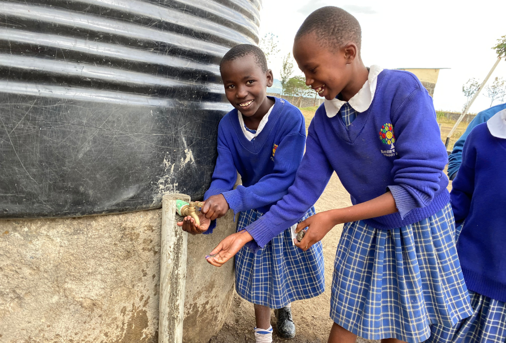 Two smiling schoolgirls in blue checkered uniforms use a water tap attached to a large black water tank outside, with one girl holding her hand under the tap and the other turning the tap handle.