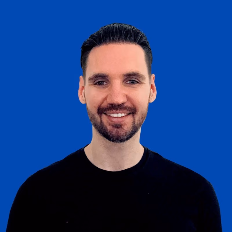 Smiling man with dark hair and beard wearing a black shirt against a solid blue background.