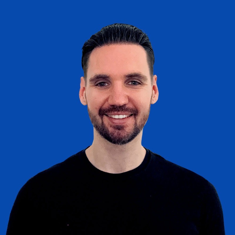 Smiling man with dark hair and beard wearing a black shirt against a solid blue background.