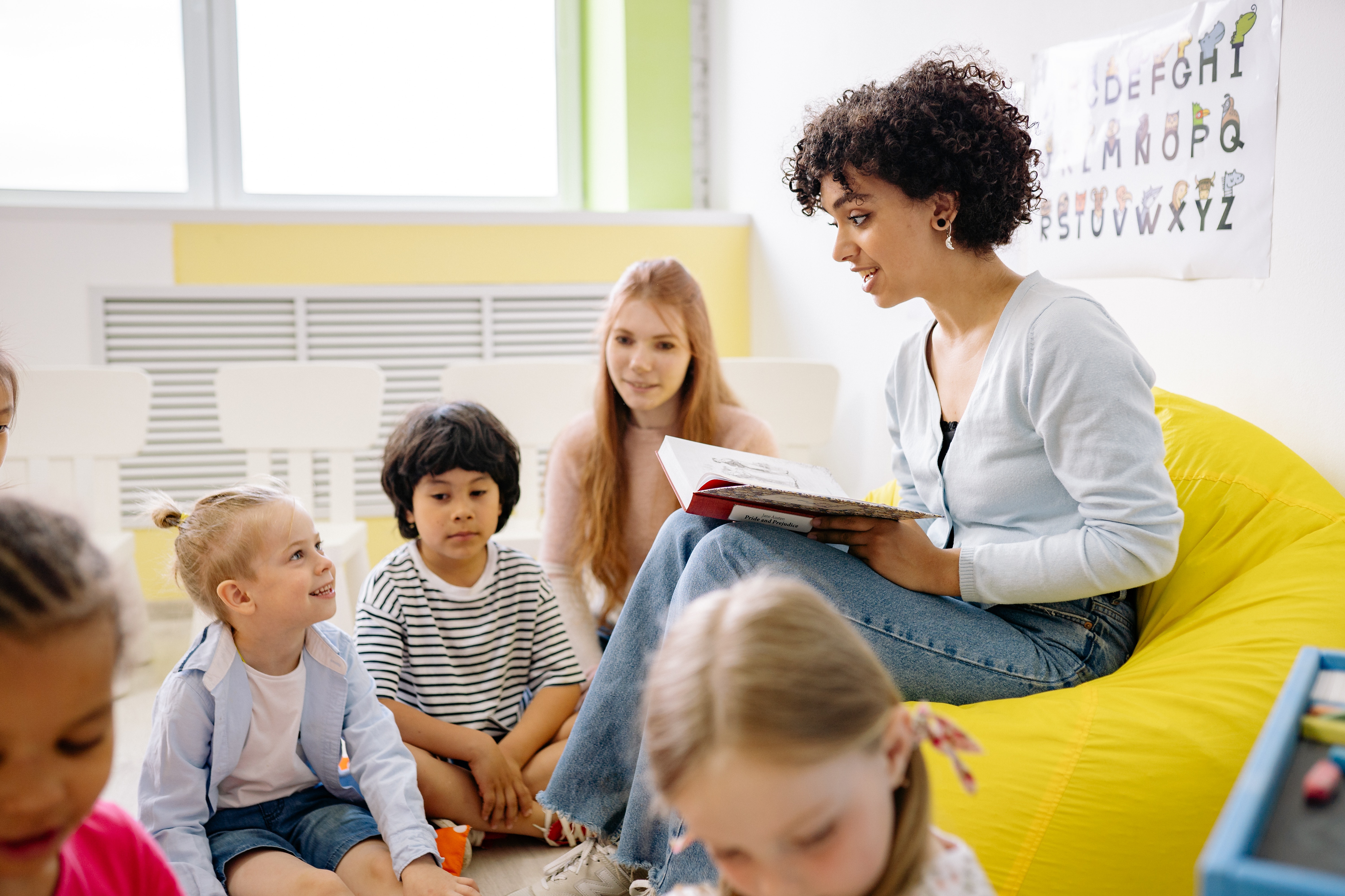 Female teacher sitting in a bright yeloow chair reading a book to students circled around her on a rug.