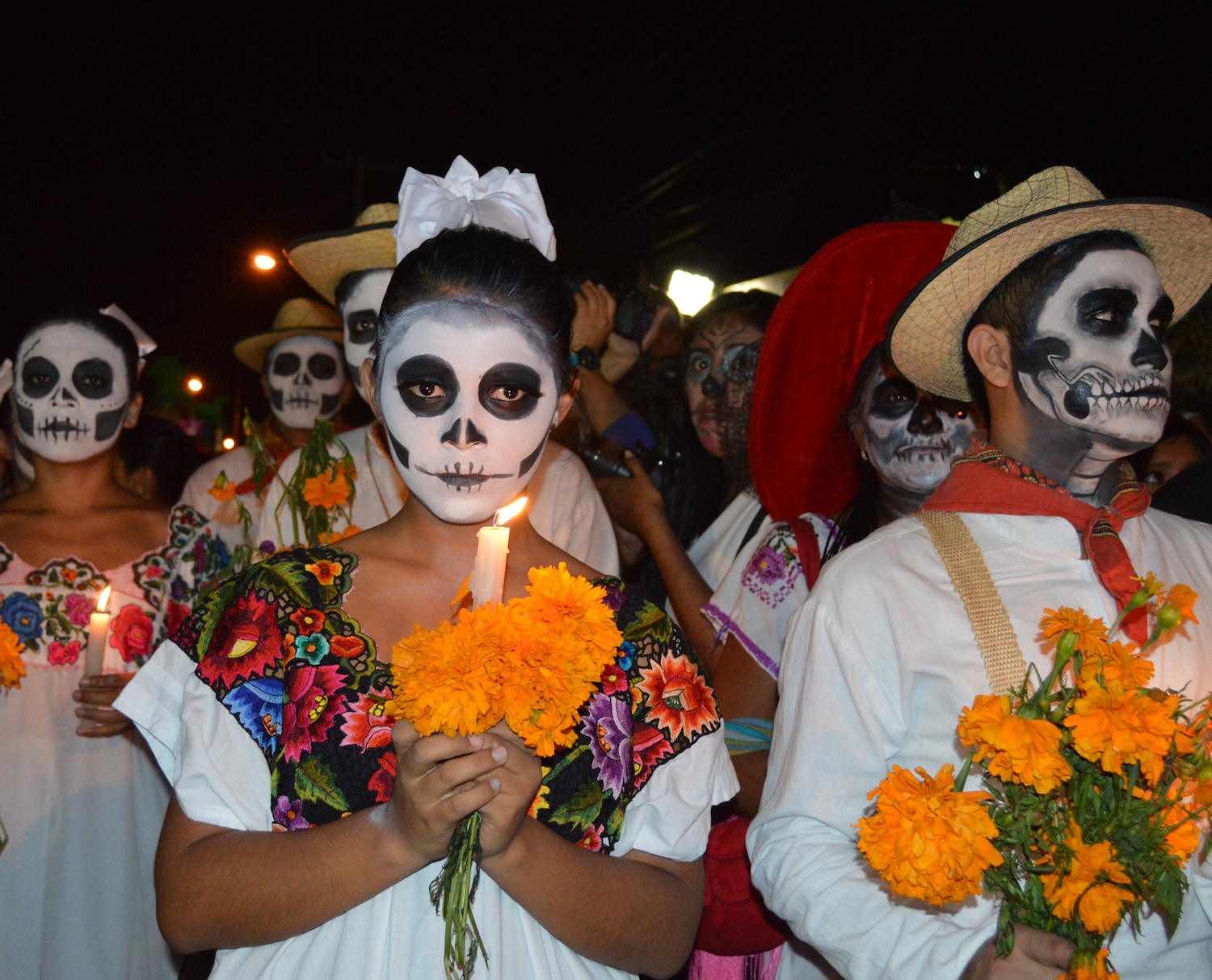 People dressed in traditional Mexican clothing with faces painted as skulls, holding marigold flowers and candles during a nighttime Día de los Muertos (Day of the Dead) celebration.