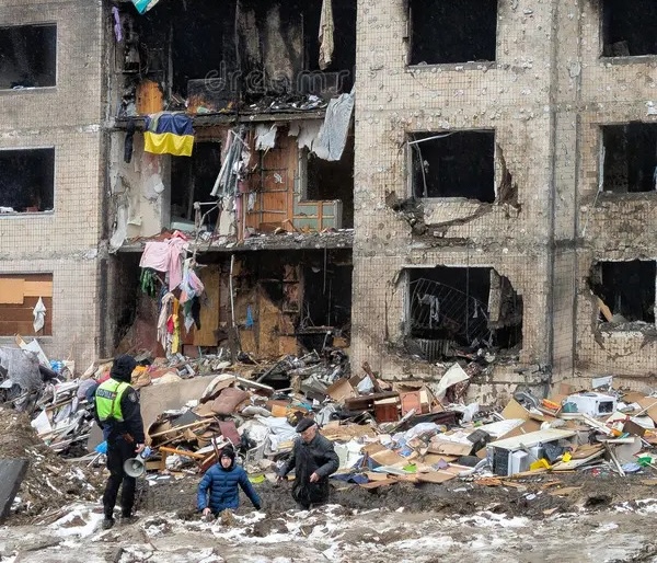 A damaged multi-story apartment building with broken windows and collapsed walls. Debris covers the ground in front of the building, where three people in winter clothing are standing and kneeling among the rubble. A flag hangs from a damaged balcony.