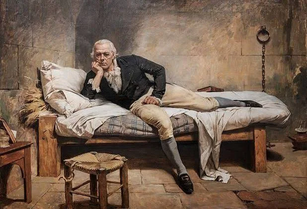 A painting of an older man reclining on a narrow wooden bed in a sparse room. He is dressed in historical clothing—light-colored trousers, stockings, and a dark jacket—and props his head on one hand while looking outward with a contemplative, weary expression. The bedding is rumpled, and a small stool and simple furnishings surround the bed. The muted tones and bare walls give the scene a somber, introspective mood