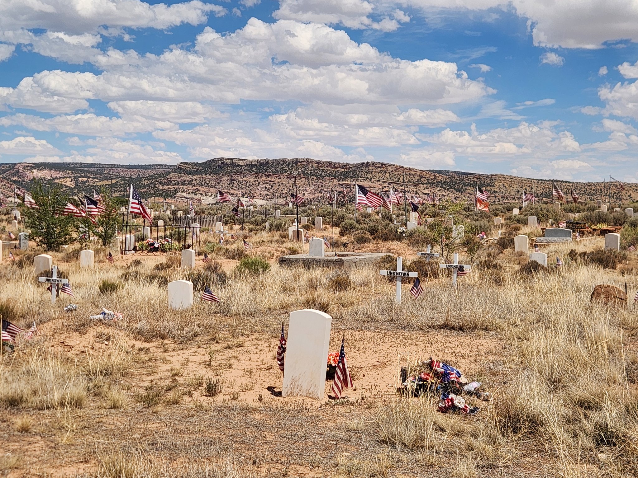 A wide view of a rural cemetery in a dry, grassy landscape, with white headstones scattered across the ground, many marked with small American flags. Low desert hills rise in the background beneath a bright blue sky filled with large white clouds.