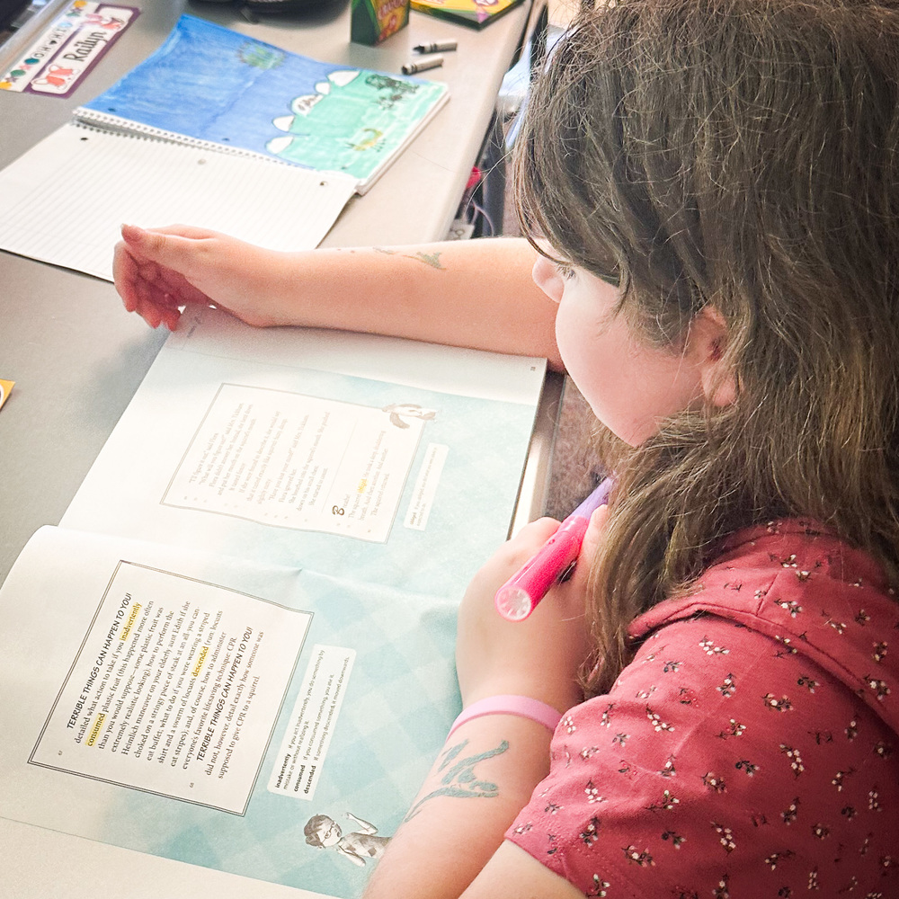 A young child with long brown hair sits at a desk working on a reading worksheet. The child holds a pink highlighter and looks down at the page. On the desk are a spiral notebook and a colorful drawing with blue sky and green landscape. The child has small temporary tattoos on their arm and is wearing a pink patterned shirt.