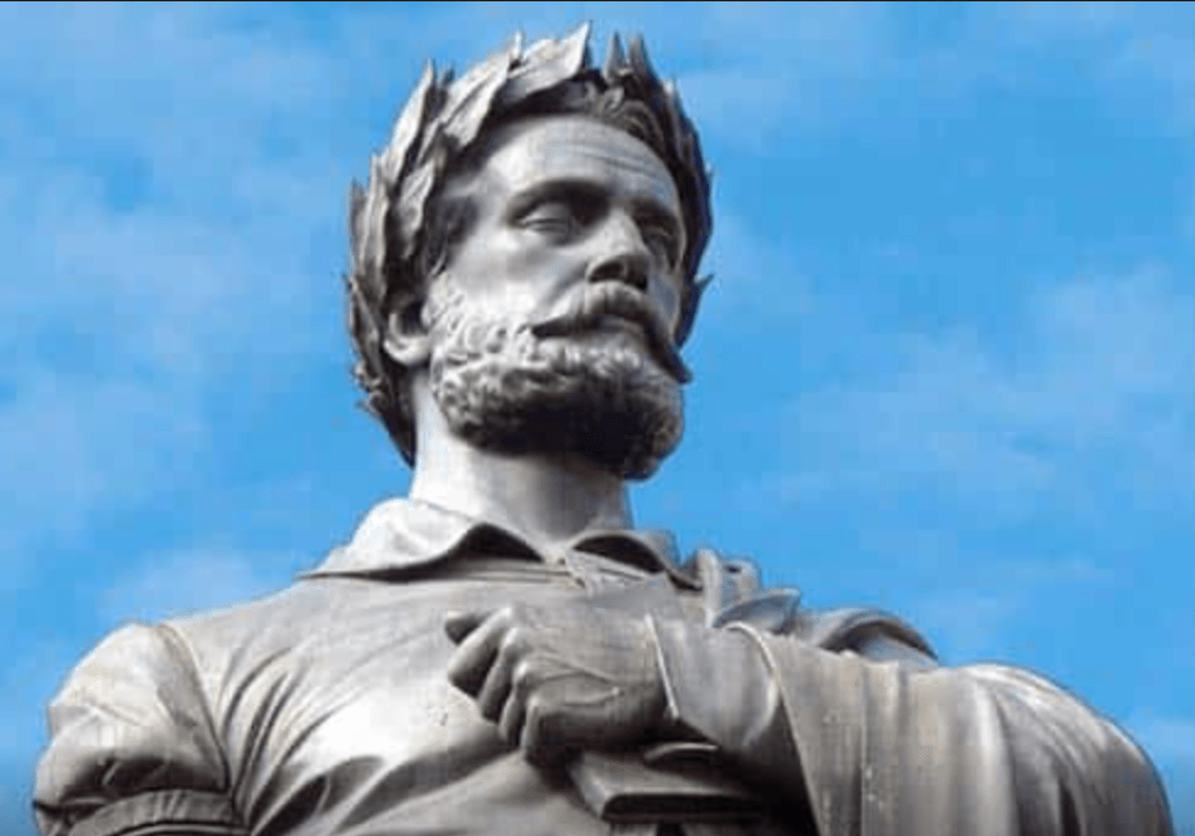 A close-up view of a stone statue of a bearded man wearing a laurel wreath, gazing slightly upward with a calm, dignified expression. His hands are positioned near his chest, holding an object that appears to be a book. The background is a clear blue sky.