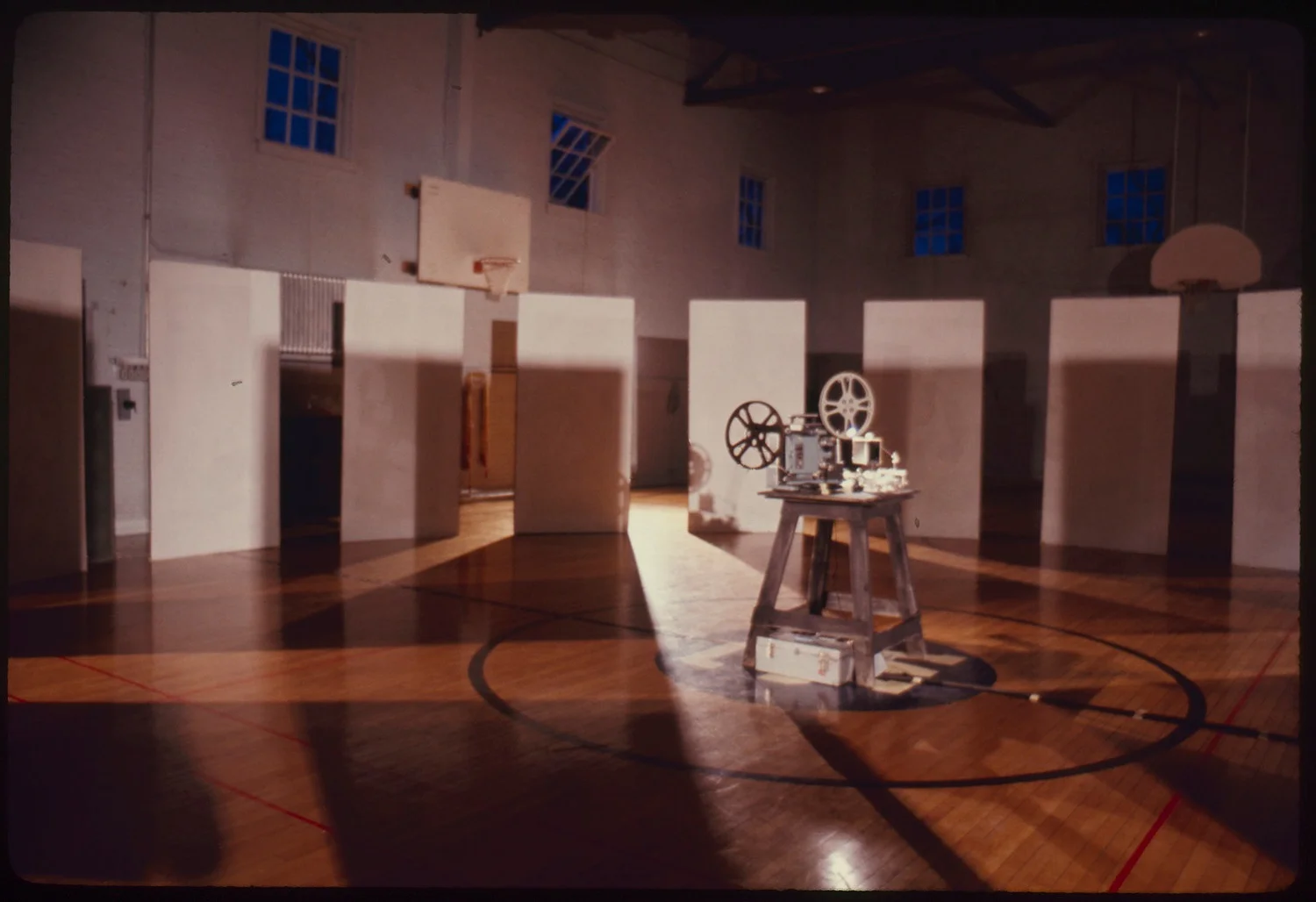 A warmly lit indoor gymnasium with a polished wooden floor and basketball hoops mounted on the walls. In the center, a small vintage film projector sits on a wooden stool, casting long shadows across the floor. Around it, several tall white panels stand upright in a semicircle, each with a doorway cut out, creating a maze-like installation. Sunlight streams in from high windows, adding a soft, cinematic glow to the scene.