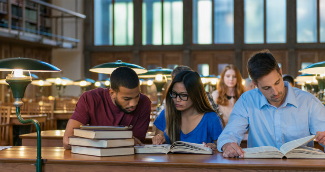 Three students sit closely together at a long wooden table in a library, studying. A stack of books is piled in front of them, and each person is focused on open textbooks. Warm desk lamps line the table, and tall windows and bookshelves fill the background, creating a quiet, academic atmosphere.