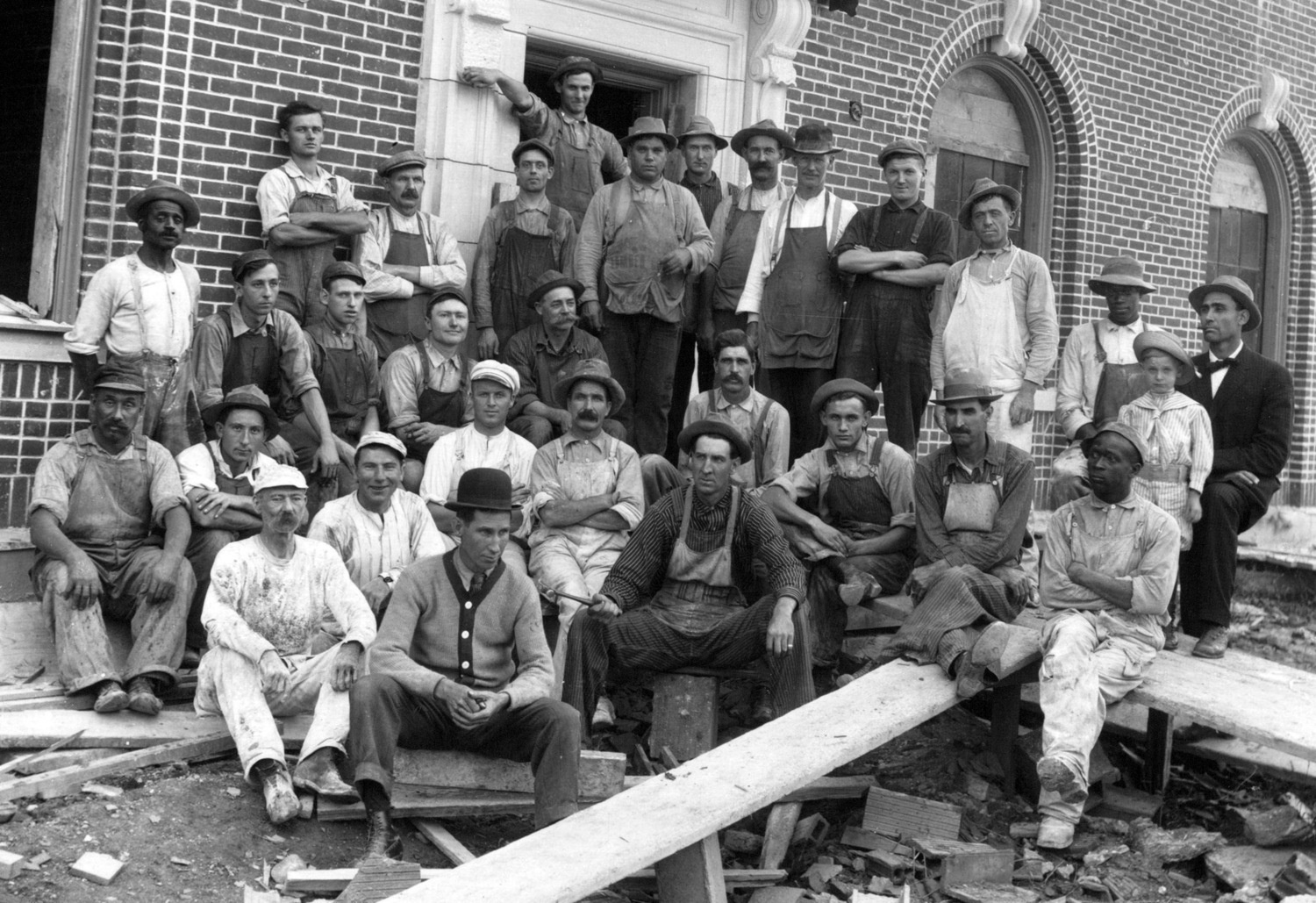 Black-and-white historical photograph of a large group of men, mostly construction or factory workers, gathered outside a brick building. They wear work clothes such as overalls, aprons, and caps, and many have mustaches. Some are seated on wooden planks and steps while others stand behind them. A few individuals appear to be Black, while most are white, reflecting a diverse workforce for the time. On the right side, a well-dressed man in a suit sits next to a young boy in a cap and striped shirt. The building behind them has arched doorways and decorative brickwork. Debris, boards, and tools are scattered in the foreground, suggesting an active worksite.