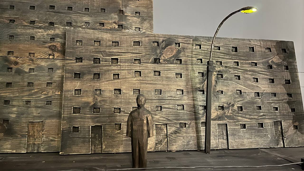 Wooden sculpture depicting a faceless human figure standing in front of dark, stained wood panels carved with square windows to resemble brutalist apartment buildings, illuminated by a single miniature curved streetlight on the right