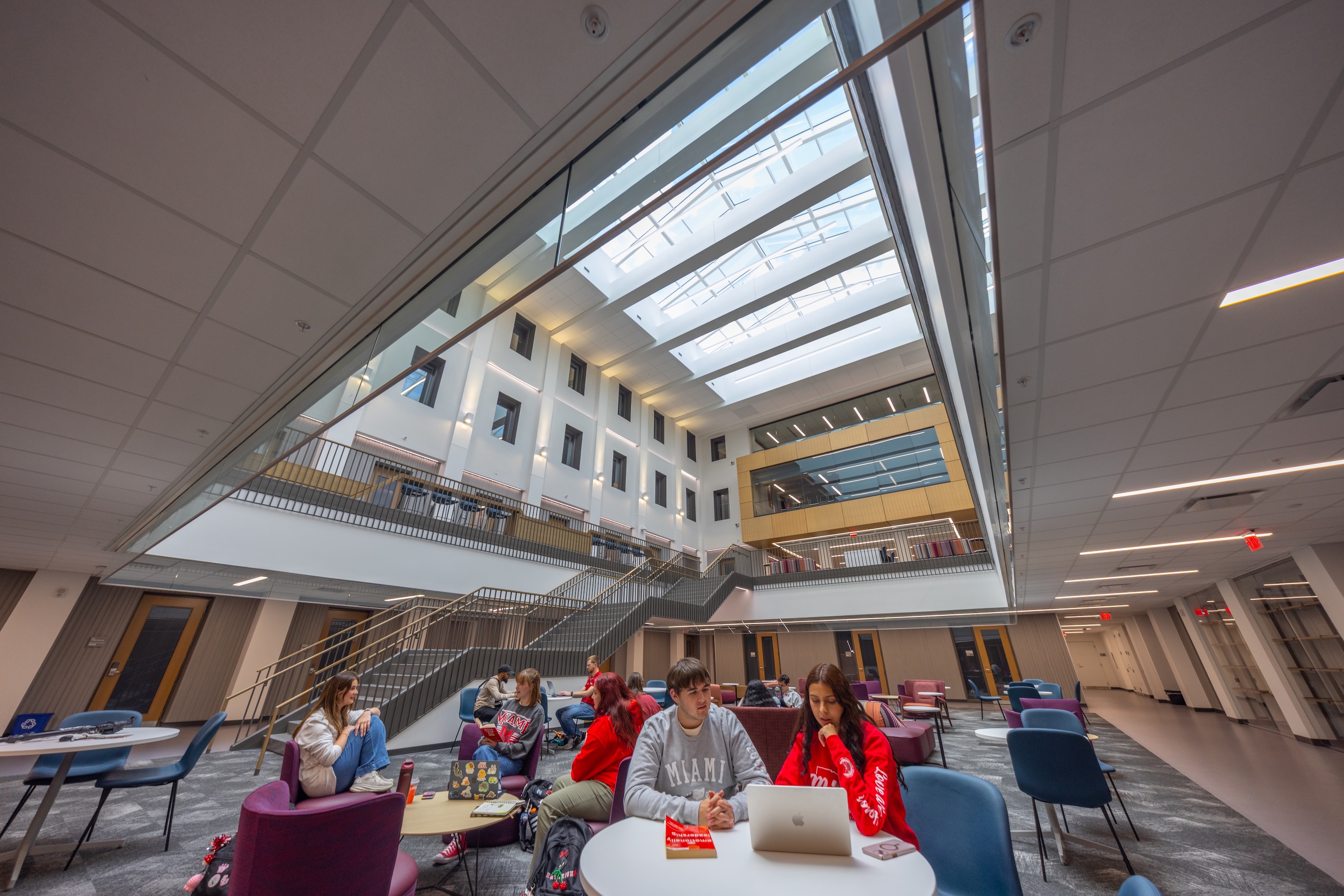 College students studying and chatting in a bright, modern multi-story atrium under a large skylight.