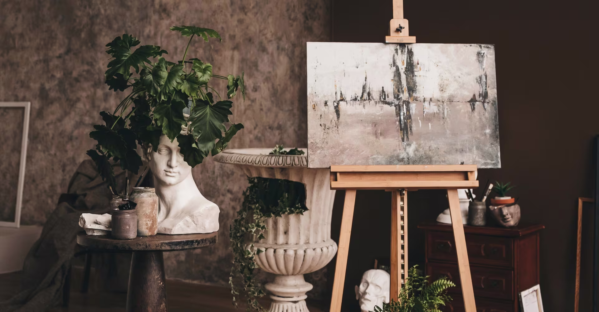 A moody, earth-toned artist studio featuring a wooden easel with an abstract canvas painting alongside classical plaster busts, lush green houseplants, and vintage stone urns.
