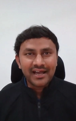 Man with short black hair wearing a black jacket, sitting against a plain white wall.