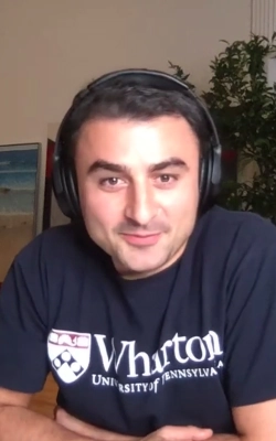 Young man with glasses and dark hair wearing a light pink shirt sitting indoors with abstract wall art in the background.