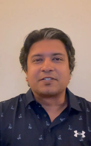 Young man with glasses and dark hair wearing a light pink shirt sitting indoors with abstract wall art in the background.
