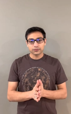 Young man with glasses and dark hair wearing a light pink shirt sitting indoors with abstract wall art in the background.