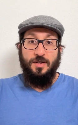 Young man with glasses and dark hair wearing a light pink shirt sitting indoors with abstract wall art in the background.