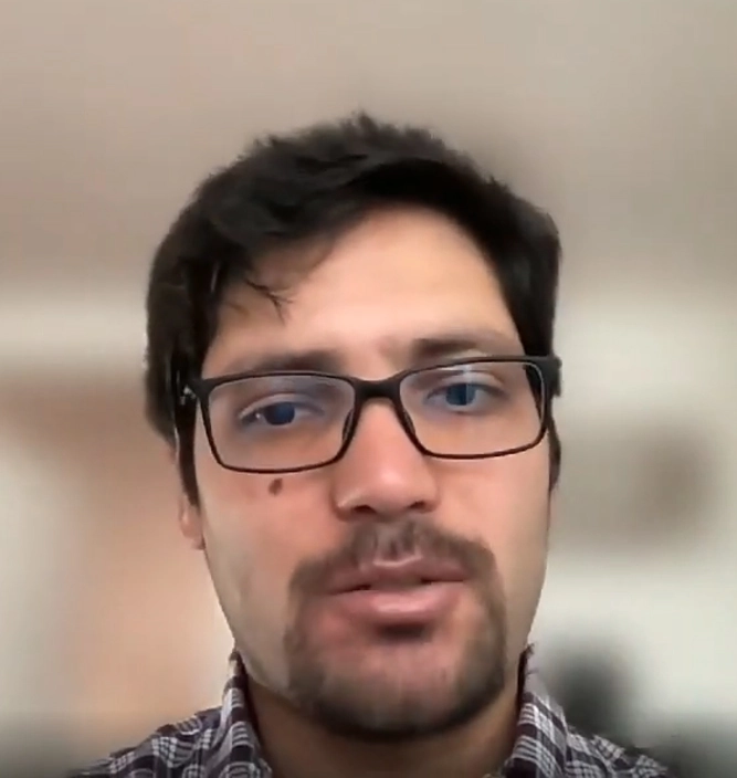 Young man with glasses and dark hair wearing a light pink shirt sitting indoors with abstract wall art in the background.
