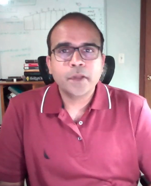 Young man with glasses and dark hair wearing a light pink shirt sitting indoors with abstract wall art in the background.