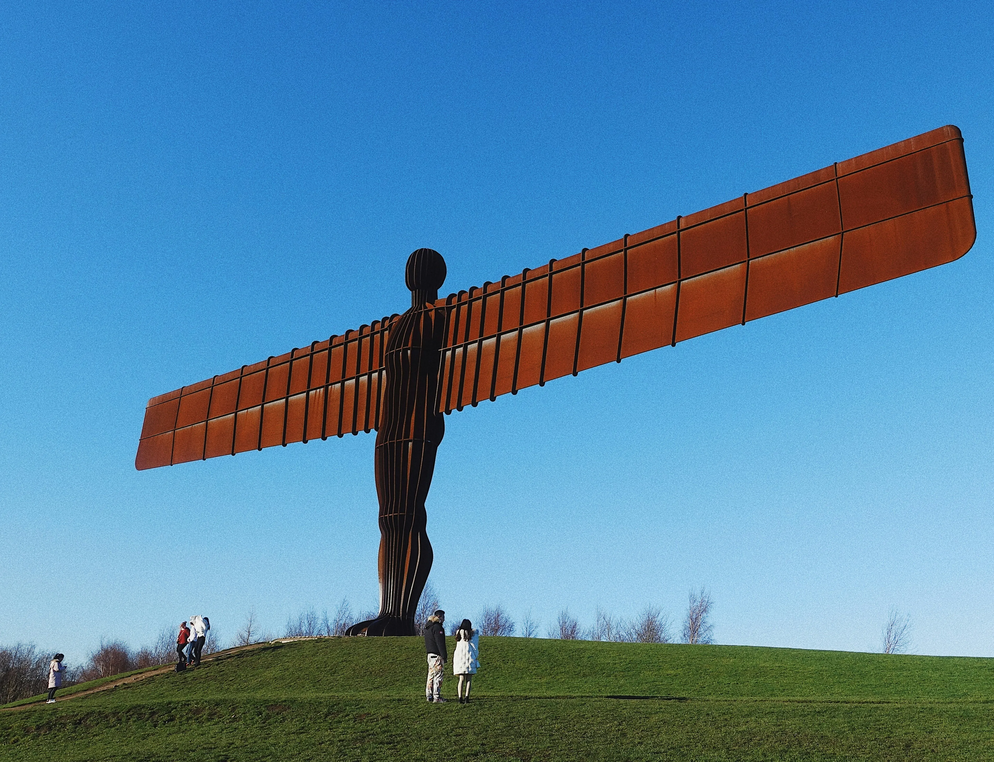 The Angel of the North sculpture stands on a grassy hill under a clear blue sky with several people nearby for scale.