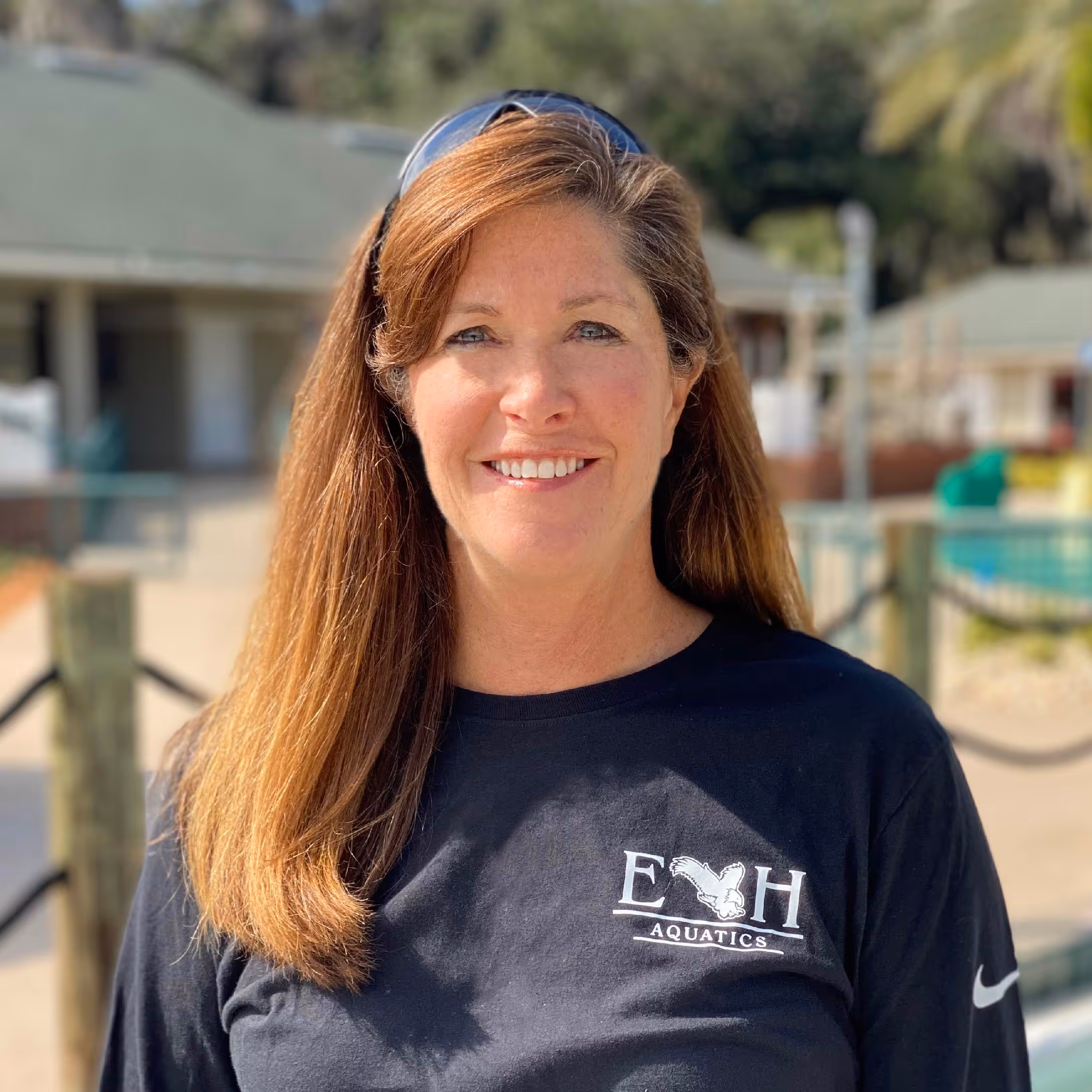 Smiling woman with long brown hair wearing a black E H Aquatics shirt and sunglasses on her head outdoors.