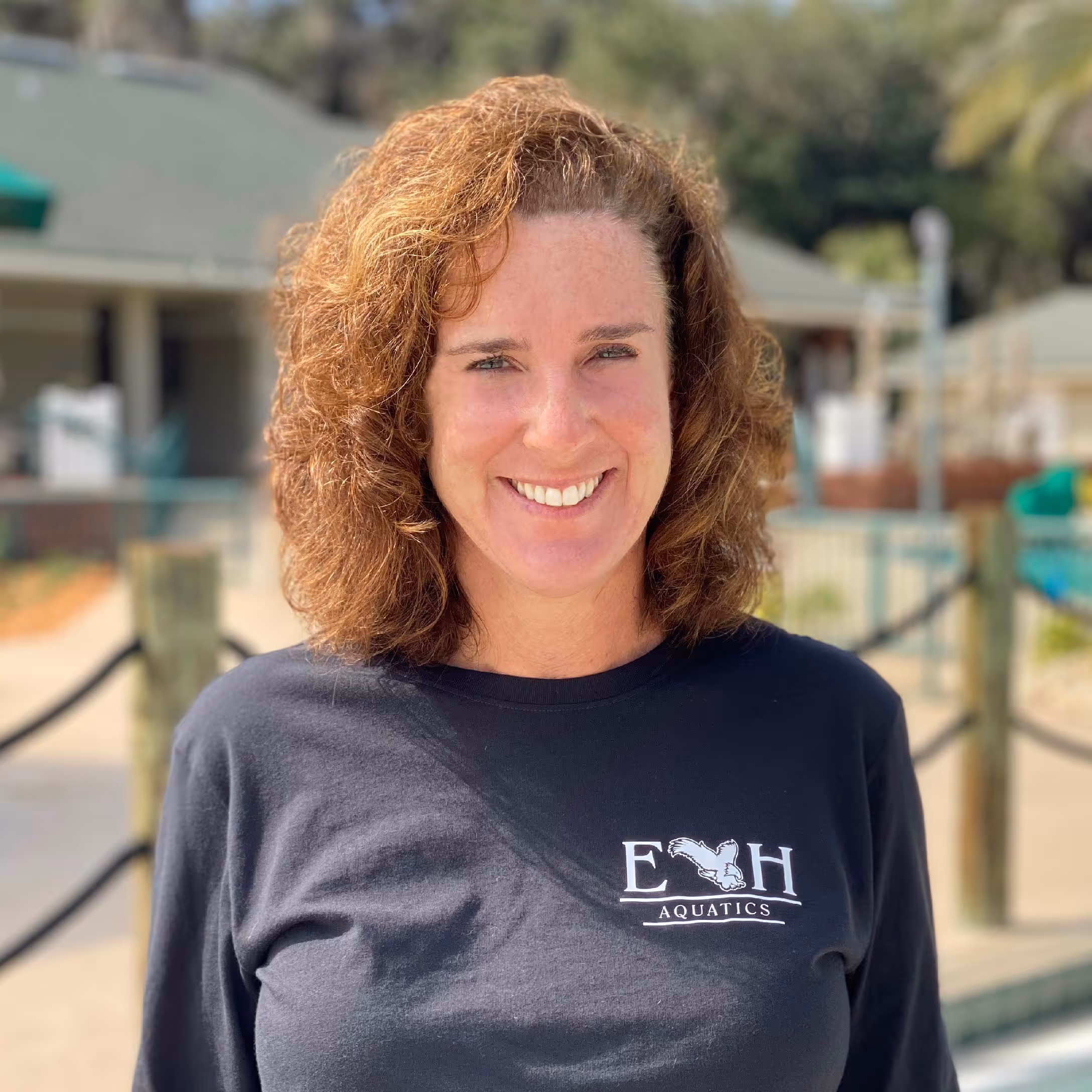 Smiling woman with curly brown hair wearing a black E H Aquatics shirt outdoors.