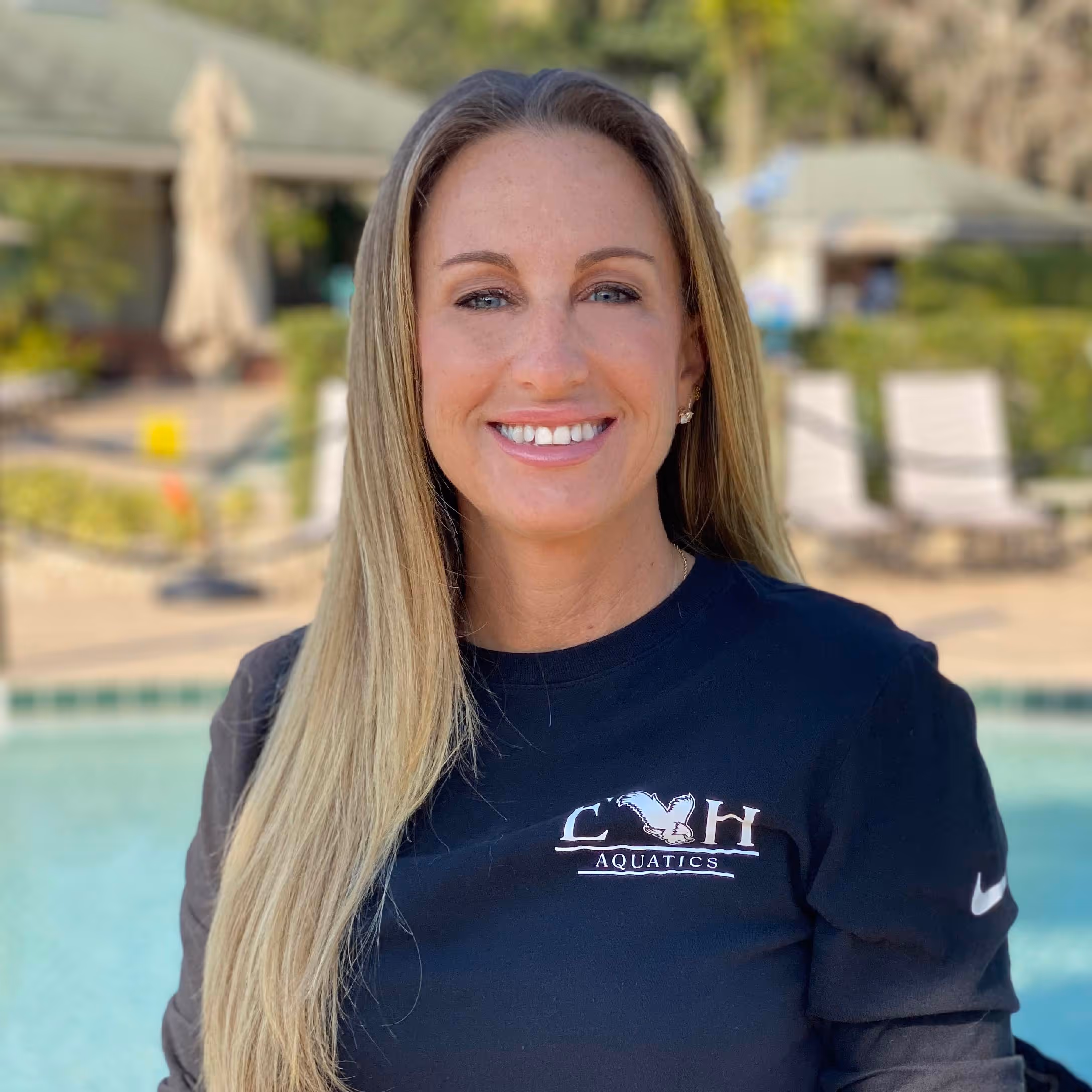 Smiling woman with long blonde hair wearing a black CVH Aquatics shirt, standing outdoors near a pool.