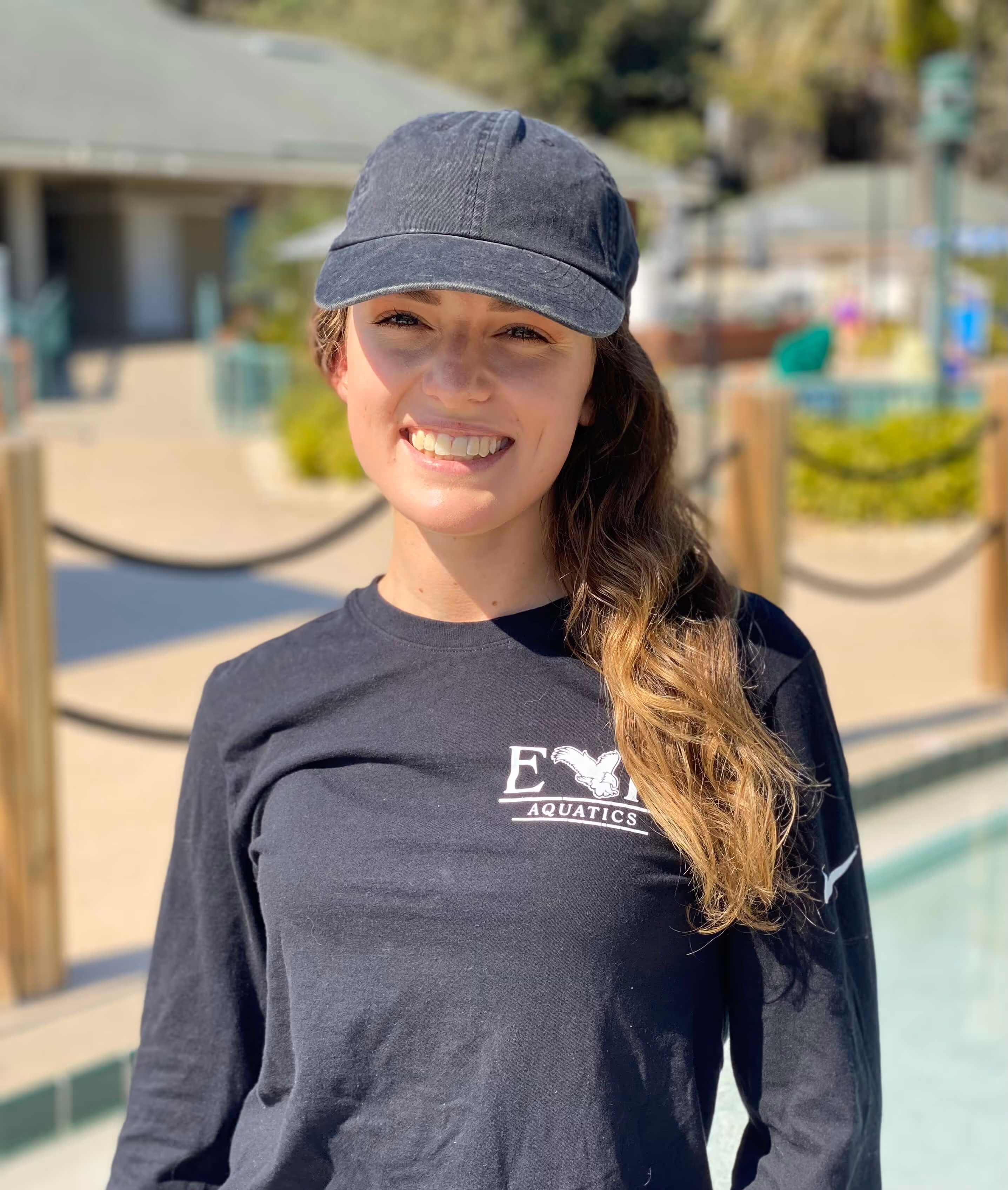 Smiling woman wearing a black cap and black long-sleeve shirt with 'EYY Aquatics' logo standing outdoors near a pool.