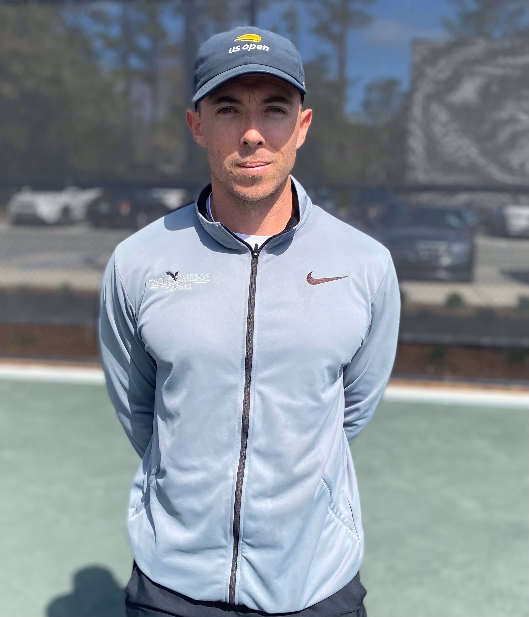 Man in a light gray zip-up jacket and US Open cap standing on a tennis court with arms behind his back.