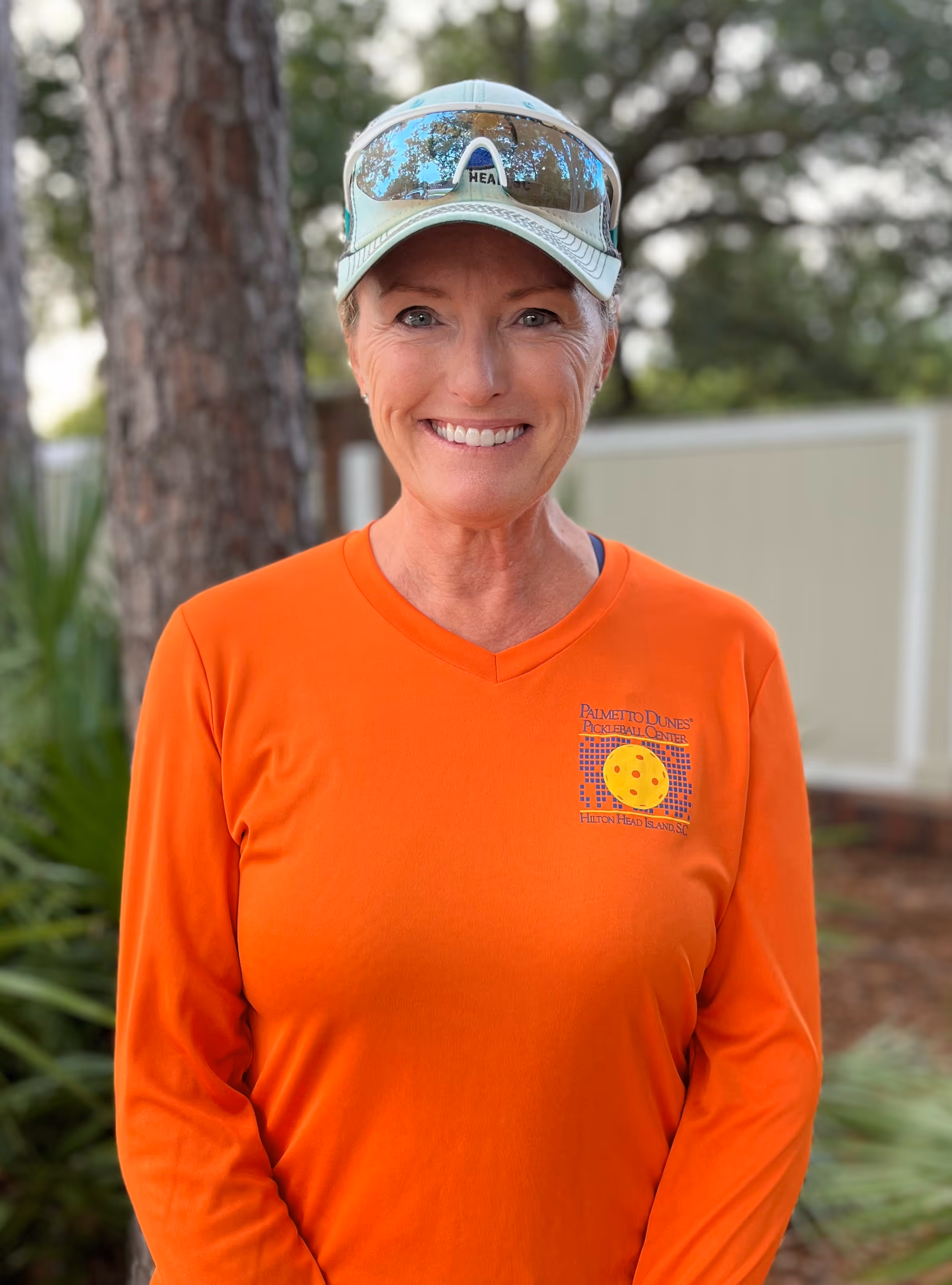 Smiling woman wearing an orange long-sleeve shirt and baseball cap standing outdoors with greenery and a tree behind her.
